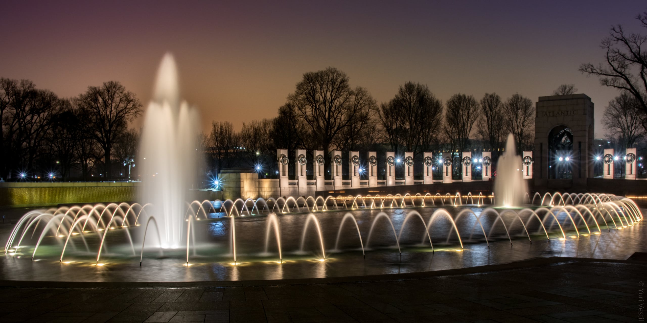 The National World War II Memorial at night. The area is well-lit, revealing a series of bronze plaques detailing various World War II veteran accounts.