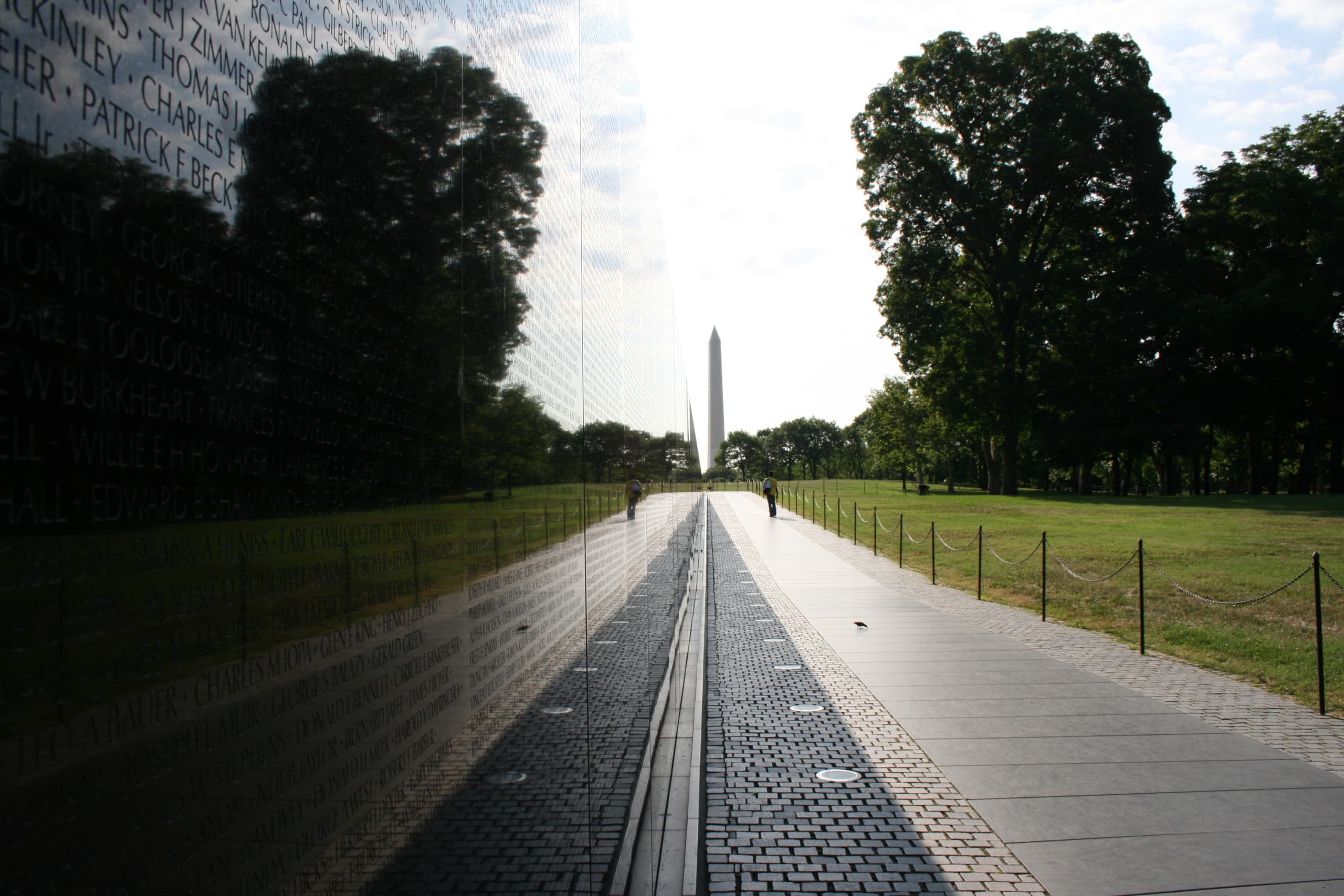 The Vietnam Veterans Memorial Wall is a large granite wall engraved with the names of 58,318 Americans who lost their lives during the Vietnam War. The Washington Monument stands in the distant background.